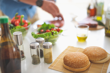 Close up of hamburger bun with sesame seeds, bottles with cumin, ground cloves and pepper on table. Vegetables and male hands cooking meat on blurred background