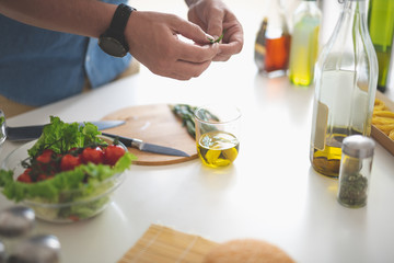 Close up of male hands adding rosemary to glass of olive oil. Bowl with fresh vegetables, bottle, spices and cutting board on white kitchen table
