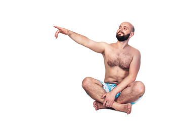 The cheerful young guy in shorts is sitting on the floor and pointing at copy space