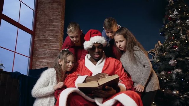 Cute santa claus in funny hat reading book with kids.