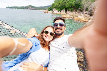 Fototapeta premium Young beautiful happy smiling funny couple man and woman best friends on a hammock on vacation makes selfie on a smartphone against the background of the sea
