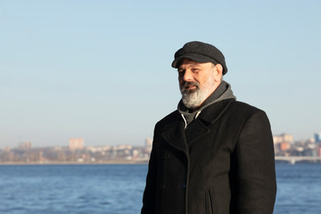 Portrait of a man with a gray beard in a black jacket and cap against the background of the city.