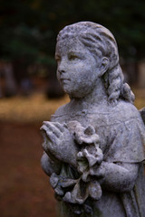 Fototapeta na wymiar Partial View of Victorian Statuary, a Girl's Face, Shoulders, and Hands in Prayer Position Holding a Wreath with Out of Focus Foliage and Trees in Background