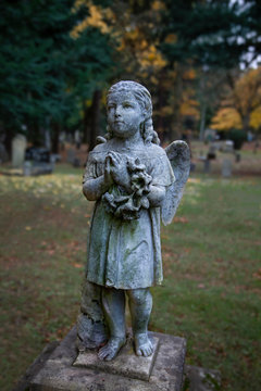 Full View of Victorian Statuary Covered in Green Moss, a Girl with Angels' Wings, Hands in Prayer Position Holding a Wreath with Out of Focus Cemetery in Background in Vertical Format