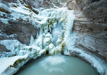 A half frozen waterfall flows into a green pool surrounded by huge ice masses from a cold winter