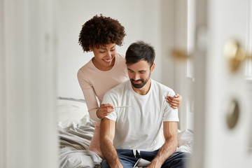 Glad dark skinned woman shows home pregancy home result to boyfriend, have smiles on faces, dressed...