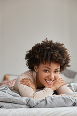 Vertical shot of satisfied pleasant looking lady with toothy smile curly hair, lies on stomach in comfortable bed, has weekend, enjoys comfort and cozy domestic atmosphere. People and rest concept