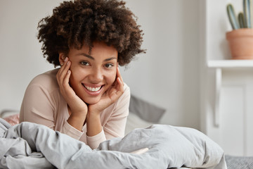 Picture of glad black young lady touches cheek, keeps both hands under chin, has Afro hairstyle, wears casual clothing, poses on bed, looks happily. People, relaxation and lifestyle concept.