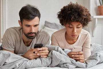 Addicted multiethnic woman and man search information in network, use modern cell phones, watch video or read weblog, have rest in bed, use wireless internet. Modern technologies and addiction