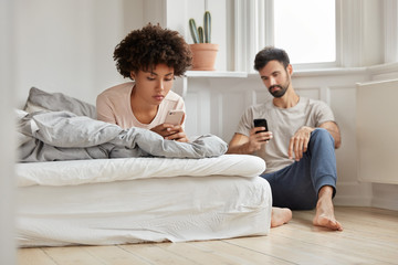 Multiethnic couple watch video via social networks, hold modern cellulars, wife lies in bed, husband sits near at floor, write text message, pose in spacious light room at home. Online communication