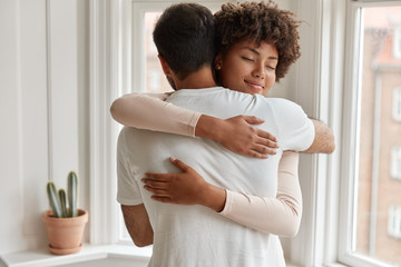 Pleased dark skinned young woman gives warm hug to her boyfriend, being pleased, pose near window, have romantic relationship, stand in cozy room. Husband and wife feel pleased and togetherness - Powered by Adobe