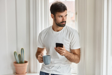 Shot of bearded Caucasian man in white t shirt, holds mobile phone and mug of coffee, installs new application, enjoys free internet, focused aside, orders food in restaurant for dinner, drinks coffee
