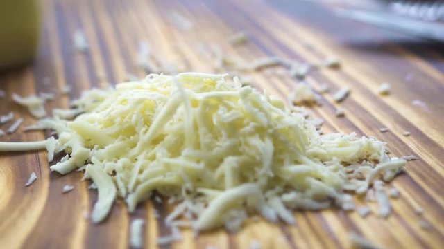 Grated cheese on wooden table close up. Process grating dairy cheese on grater on kitchen board. Ingredients for cooking pasta, pizza, salad, lasagna. Mediterranean, italian and greek cuisine.
