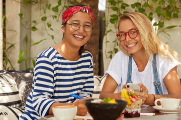 Photo of cheerful two mixed race students meet at cafe for completing common task, enjoy tasty dish, smile broadly, wear optical glasses, chat on cell phone, write exercise in notepad, drink coffee