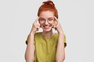 Use your brain. Happy red haired girl holds both index fingers on temples, tries to think before...