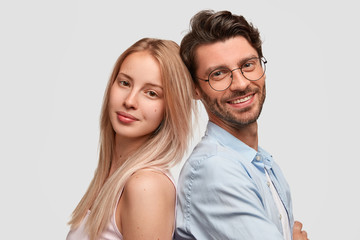 Lovely couple stand back to each other, being in good mood, isolated over white background. Pretty blonde lady, unshaven cheerful man in spectacles being partners, feel support, ready to help