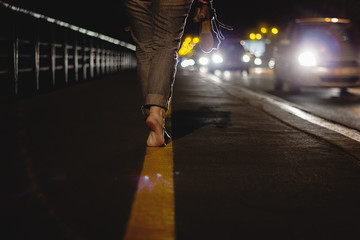 Woman walking bare foot on tip toes in night with incoming traffic, wearing her shoes in her hand