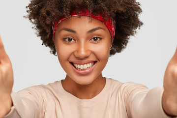 Close up shot of cheerful dark skinned girl talks with boyfriend via online video, touches camera, makes selfie with unrecognizable device, has broad smile, models over white wall, pulls hands forward