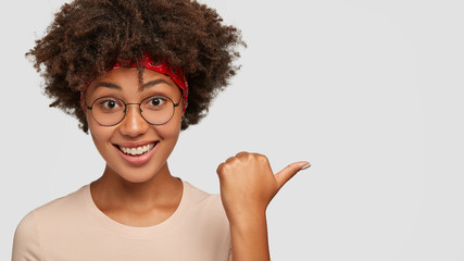 Happy black ethnic woman smiles joyfully, points with thumb aside, shows store where she bought new outfit, wears spectacles, being amuzed by something, models against white blank studio wall