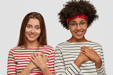 Fototapeta premium Two delighted happy multiethnic women hold hands on breast, smile joyfully and remember great moment, appreciate somebodys support, feel thankful, wears striped jumpers, isolated over white background