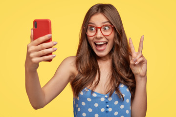 Attractive glad European woman with long hair, makes peace sign, holds mobile phone, poses for selfie, dressed in polka dot blue dress, isolated over yellow background makes blog about everyday life © WHstudio Leushin N