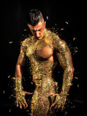 Muscular naked man covered with golden touching chin and looking down sensually on black background