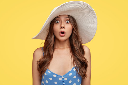 Speechless shocked woman notices something terrible and horrible, wears white beach hat, blue dress, keeps jaw dropped from fear, isolated over yellow background, widens eyes with amazement.