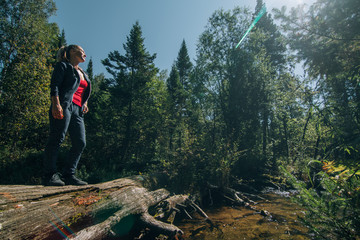 Fototapeta premium Beautiful girl stand on log and chilling after travel. Woman traveler in summer passes on wooden bridge in background of forest. Bridged lies on the river. Model of caucasian is chill and happy.