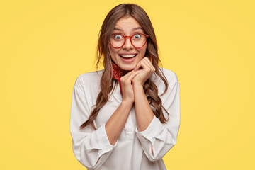 Pretty satisfied lady expresses good feelings, keeps hands together under chin, has happy look,...