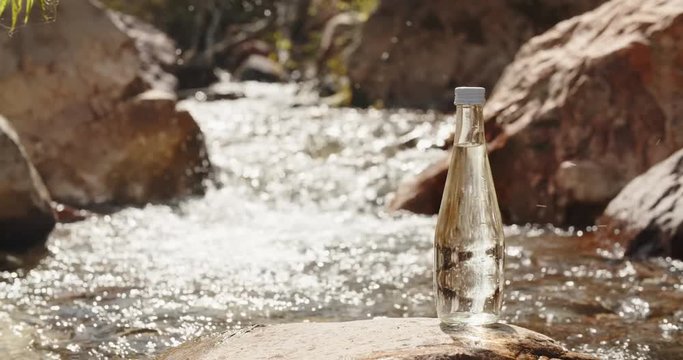 Glass bottle of water standing on rock, sunbeams shining through it, while pure river flows in stony mountains with murmuring - tranquility, harmony with nature 4k