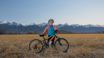 Obraz na płótnie Canvas One caucasian children walk with bike in wheat field. Little girl walking black orange cycle on background of beautiful snowy mountains. Biker stand with backpack and helmet. Mountain bike hardtail.