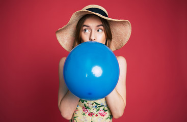 Young emotional cute blonde woman in hat is blowing blue balloon on pink background and looks in camera