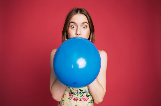 Young emotional cute blonde woman is blowing blue balloon on pink background and looks in camera