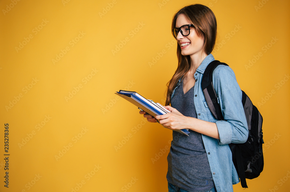 Wall mural Happy and excited cute young student girl portrait in glasses with backpack isolated in studio