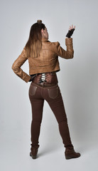 full length portrait of brunette  girl wearing brown leather steampunk outfit. standing pose with back to the camera,  on grey studio background.
