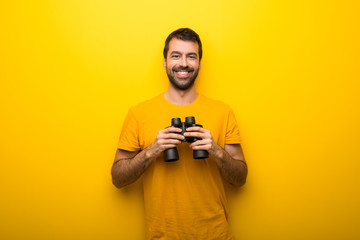 Man on isolated vibrant yellow color and looking for something in the distance with binoculars