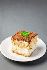 Piece of traditional italian Tiramisu dessert cake on white plate, decorated with cocoa powder and mint, on gray background, vertical, copy space