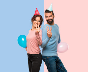 couple with balloons and birthday hats happy and counting three with fingers on pink and blue background