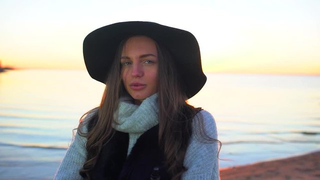 Portrait of young beautiful woman in hat at sunset smokes e-cigarette outdoors in summer. Vape. Vapping. Sea ocean beach. The girl smoking electronic cigarette and smokes. health hazards, cigarettes