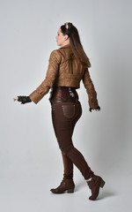 full length portrait of brunette  girl wearing brown leather steampunk outfit. standing pose with back to the camera,  on grey studio background.