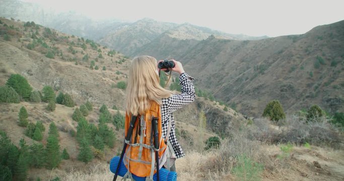 Woman blond traveler looking through a telescope on top of a mountain. 4k, slow-motion