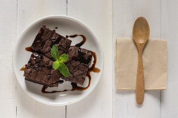 A plate of brownies with chocolate sauce on white wooden background, mint leaf on top, top view