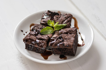 A plate of brownies with chocolate sauce on white wooden background, mint leaf on top,
