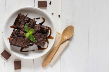 A plate of brownies with chocolate sauce on white wooden background, mint leaf on top, top view
