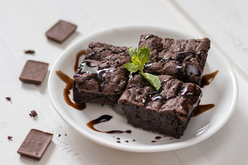 A plate of brownies with chocolate sauce on white wooden background, mint leaf on top,