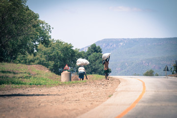 Zwei Menschen aus Afrika mit schweren Säcken auf dem Kopf laufen eine Straße entlang