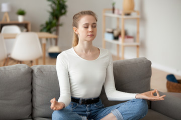 Calm girl sit in lotus position on couch practicing yoga at home, peaceful female meditating at...