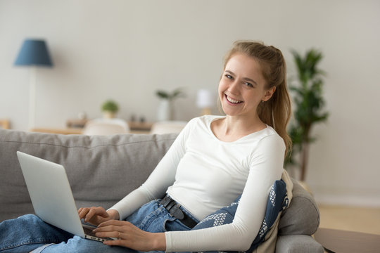 Portrait of smiling girl lying on couch typing at laptop at home, happy millennial woman sit on cozy sofa using computer indoors, excited young female look at camera working or studying online