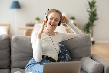 Excited girl in headset having fun at home listening to music on laptop, happy young female relax enjoying favorite tracks in earphones, millennial woman dance sitting on couch moving to the rhythm