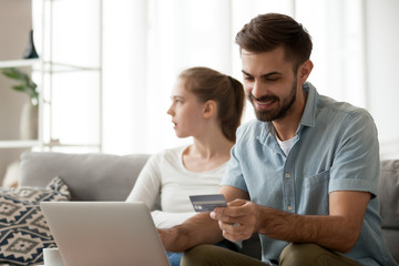 Happy millennial husband enter credit card detail making purchases on internet, dissatisfied wife sitting beside, excited man shopping online spending family money not pay attention to unhappy spouse